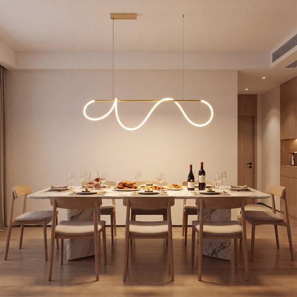 Polished-brass linear chandelier suspended above a long dining table, with flowing LED loops casting a soft glow across a warm modern room.
