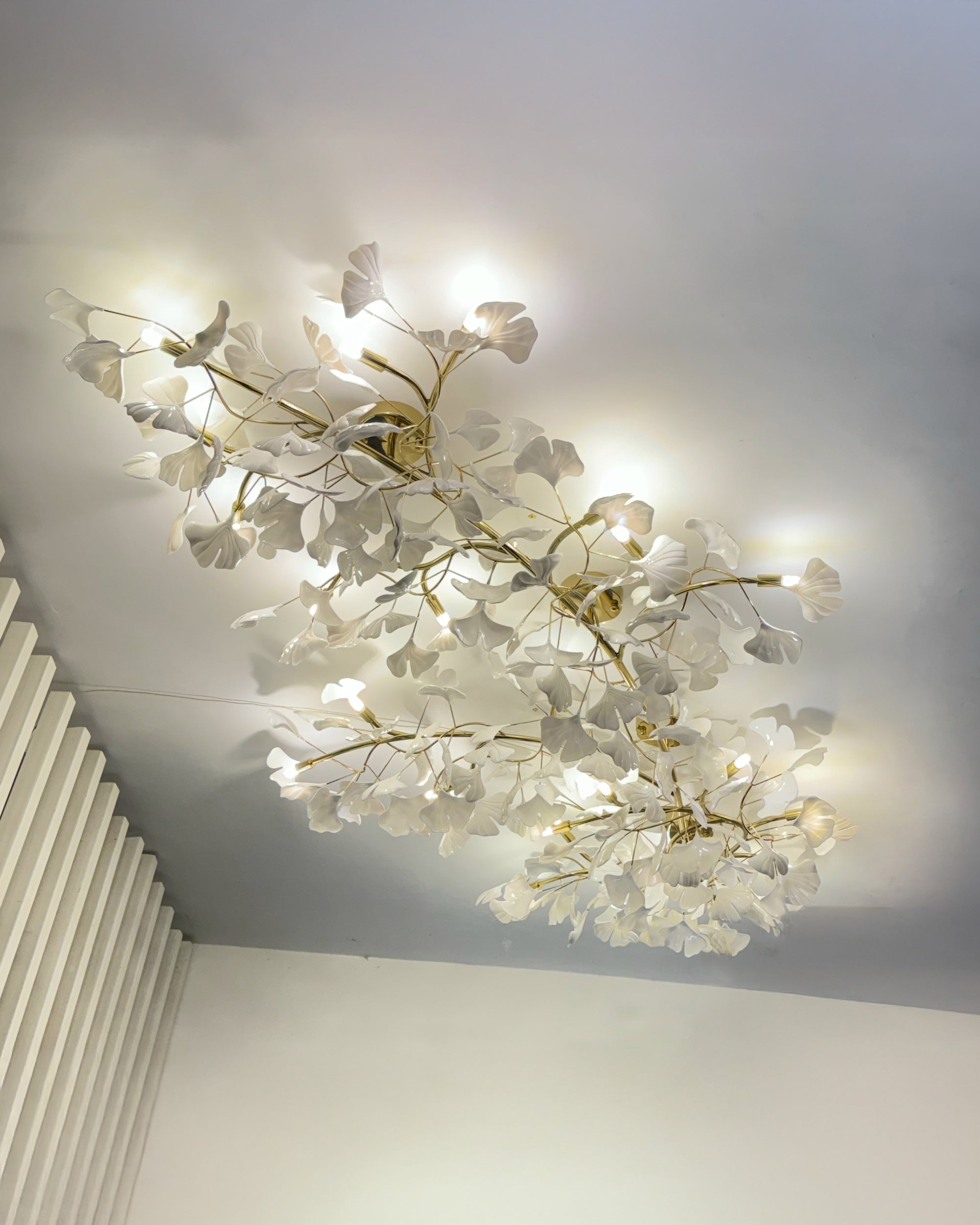 Low profile ceiling light viewed from a corner, showing white ceramic ginkgo leaves, gold branches and warm glow.
