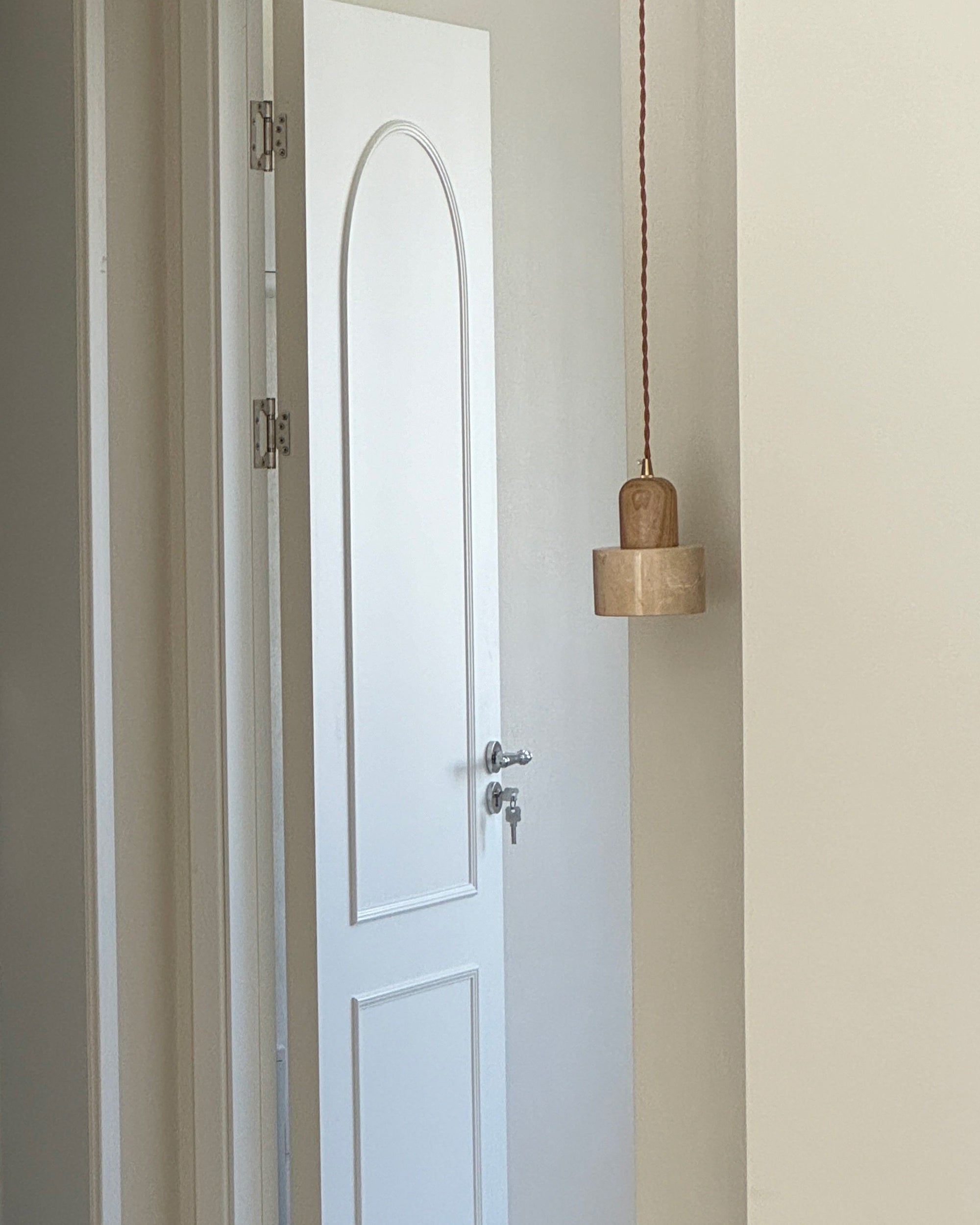 A natural pendant light with a smooth travertine cylinder and light wood top hanging beside a white doorway, shown unlit.