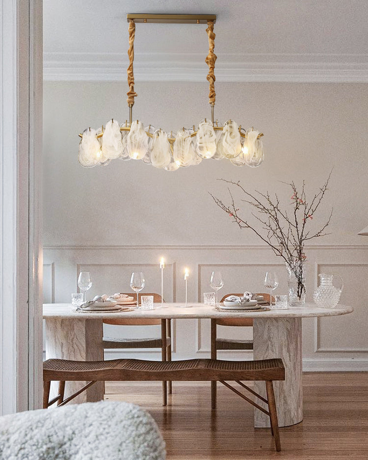 Illuminated dining room chandelier suspended above a pale stone table in a refined neutral dining room, with white-swirled glass arranged along a curved gold frame.