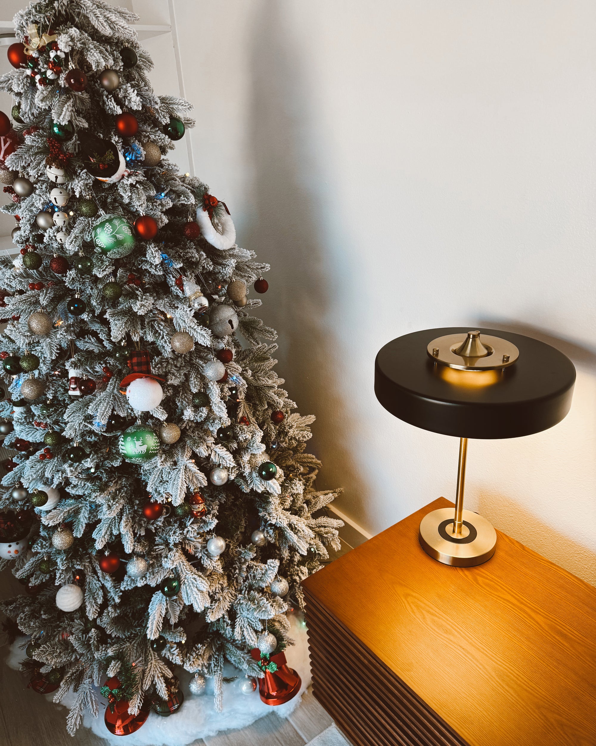 Black mid century table lamp glowing on a wooden cabinet beside a snow-dusted decorated Christmas tree.