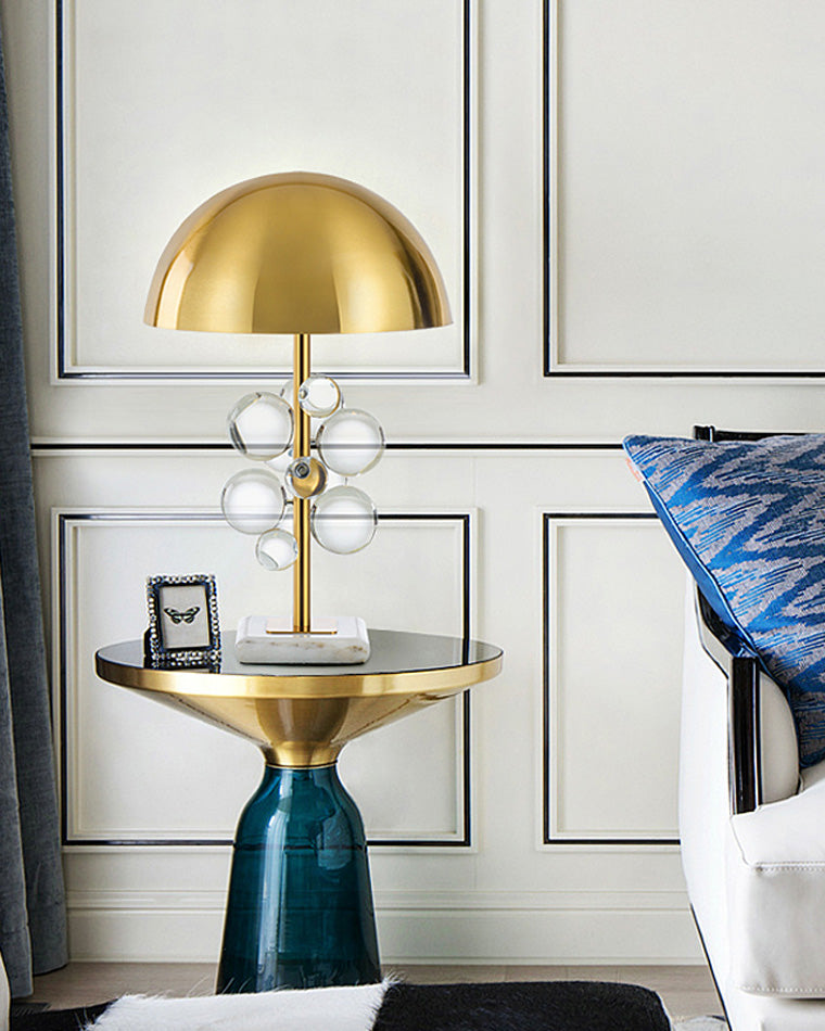 Clear brass lamp with acrylic spheres and a marble base on a blue glass side table beside a white chair.