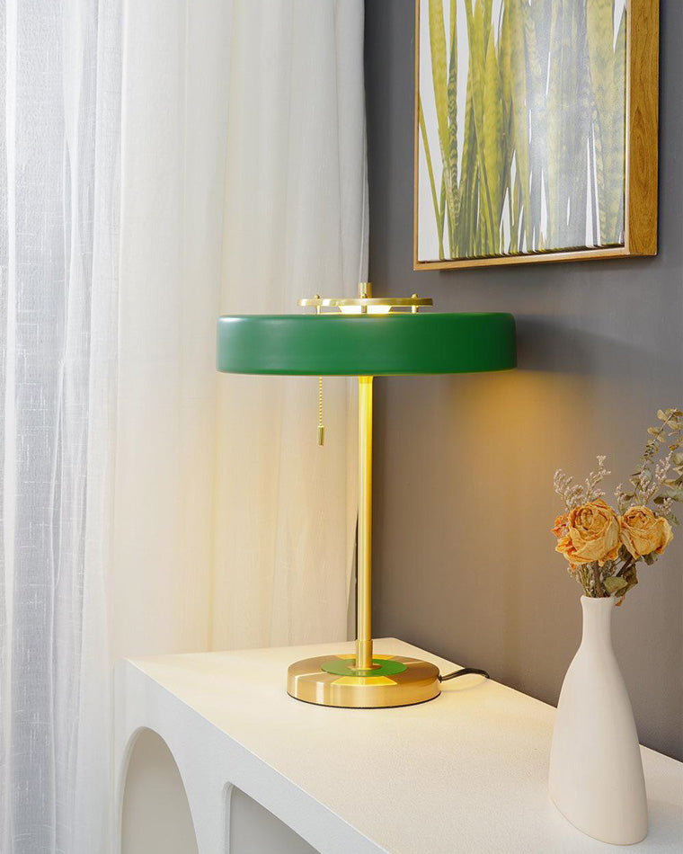 Green mid century modern table lamp glowing on a white console beside sheer curtains and a tall vase of dried flowers.
