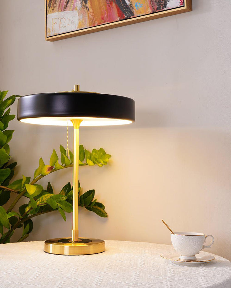 Glowing black table light with a broad round shade and gold-tone base on a covered table beside a teacup.