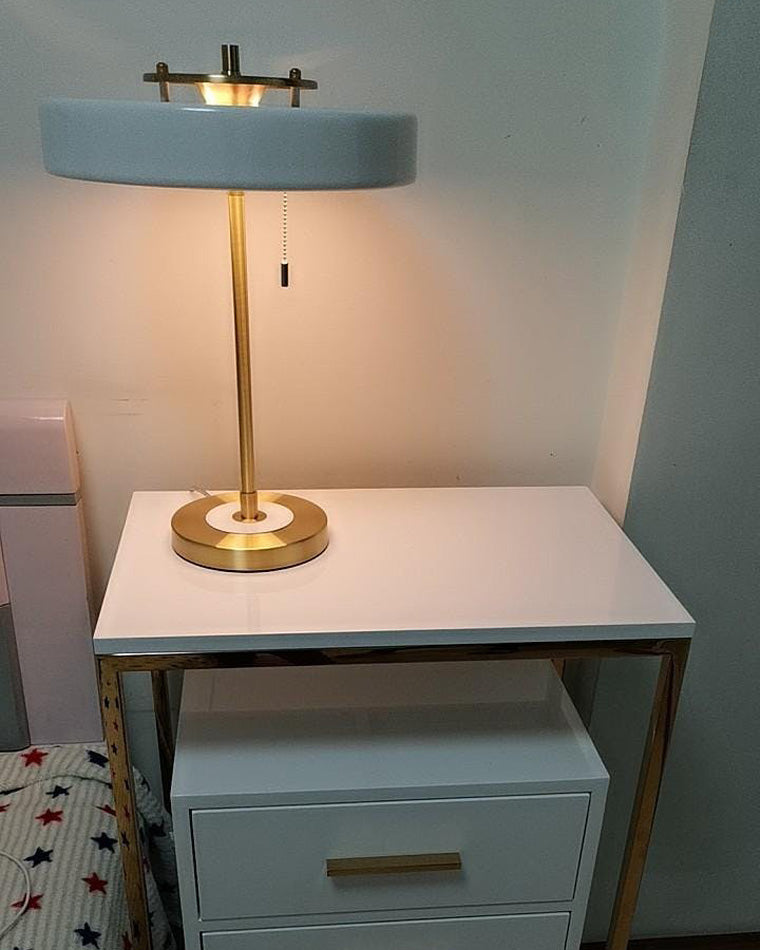 Pale blue bedside lamp casting warm downward light from a wide round shade on a white nightstand with gold trim.