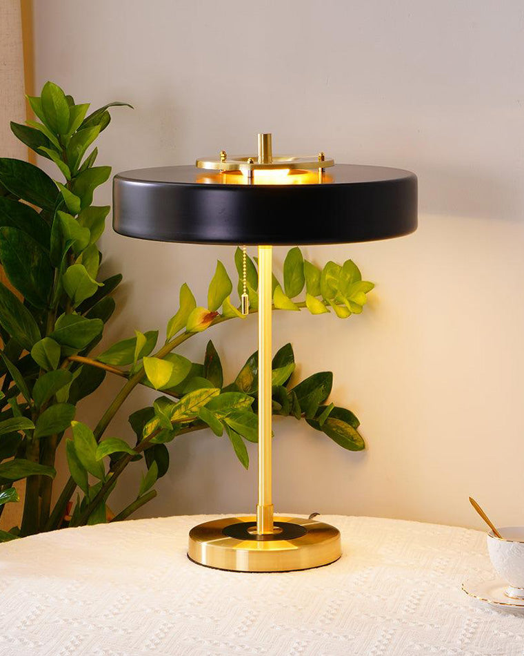 Black pull chain lamp with a glowing underside, gold-tone frame and hanging chain beside dense green foliage.