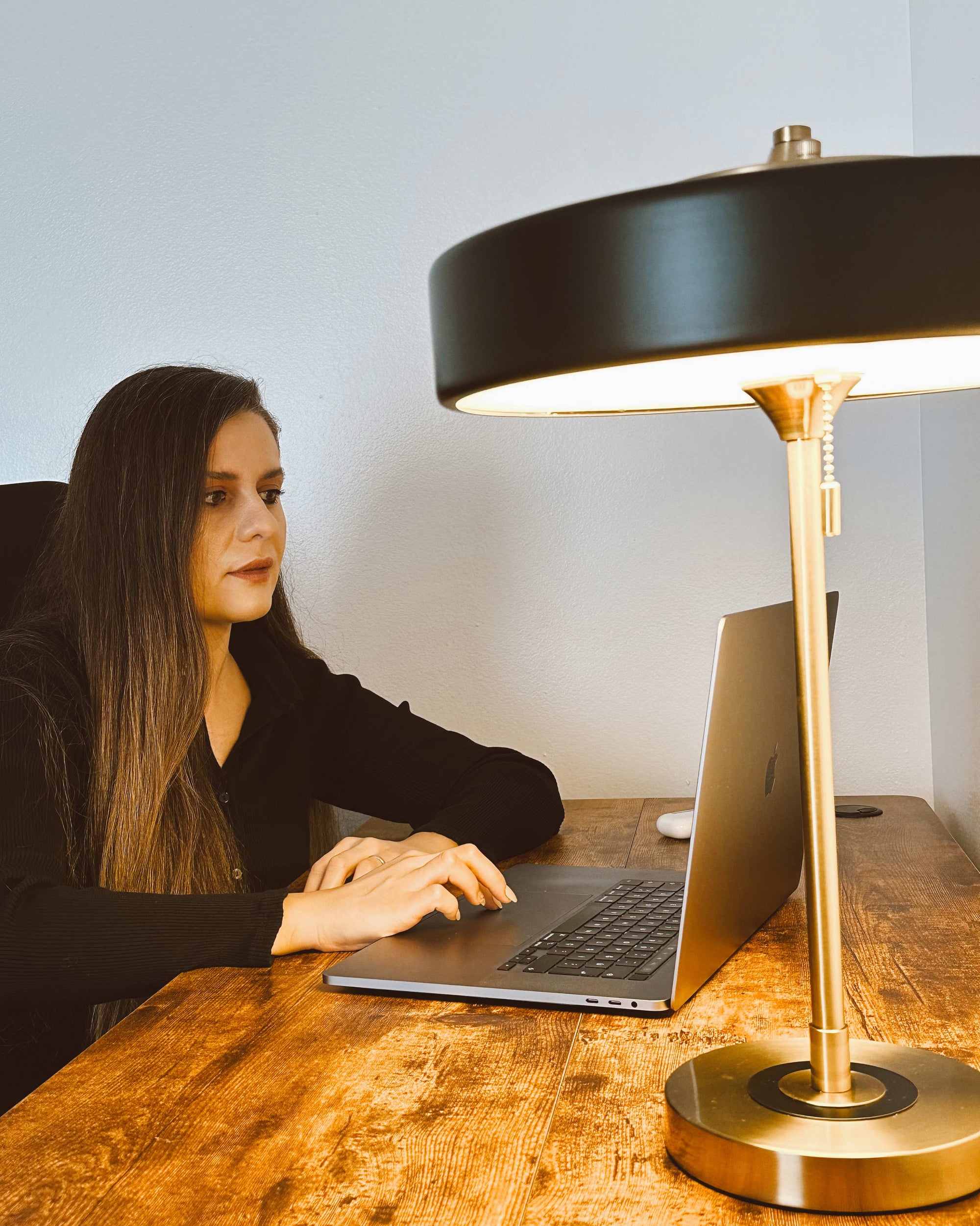 Black desk lamp with a wide metal shade lighting a wooden workspace while a woman uses a laptop.