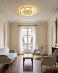 Decorative floral ceiling light centered above a traditional living room with ornate molding, pale seating and herringbone wood floors.