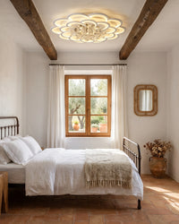 Layered flower ceiling light with crystal drops above a rustic bedroom with exposed wood beams, white bedding and terracotta flooring.