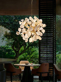 WOMO WM3353 dining room chandelier with sculpted white ceramic petals and golden brass branches glowing above a round table beside garden windows.