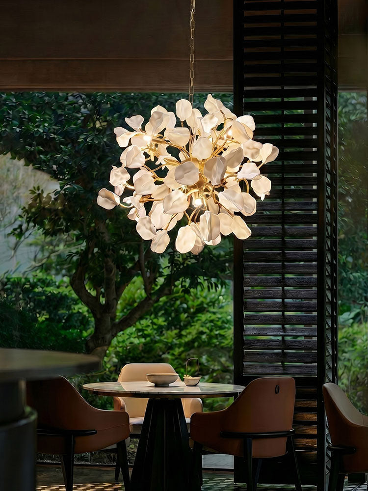 WOMO WM3353 dining room chandelier with sculpted white ceramic petals and golden brass branches glowing above a round table beside garden windows.