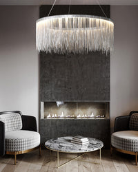 Silver waterfall chandelier shimmering above a marble coffee table and dark fireplace, lifting the moody lounge with movement and reflected light.