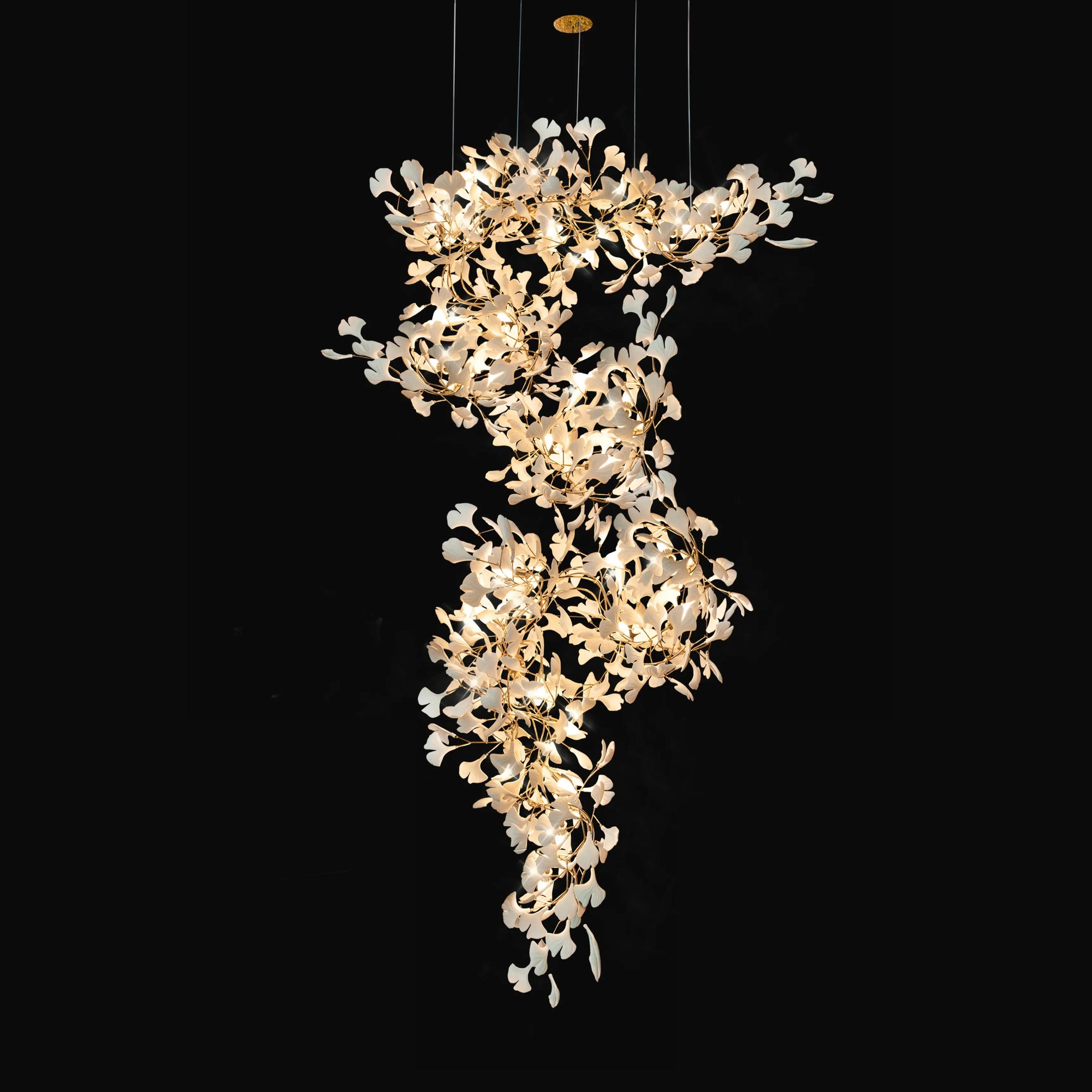 High ceiling chandelier with a long cascade of illuminated white ceramic ginkgo leaves and gold-toned branches on black.