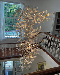 Long chandelier for staircase with white ginkgo leaves and warm bulbs, photographed from an upper landing.