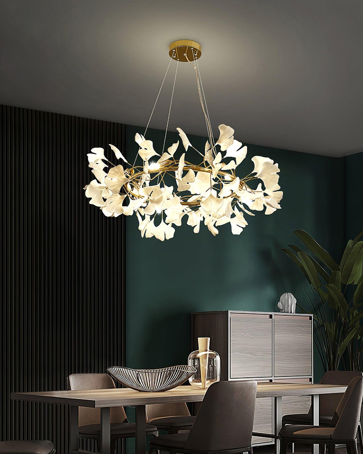 Gold-framed ceramic leaf chandelier above a dining table against deep green and black walls.