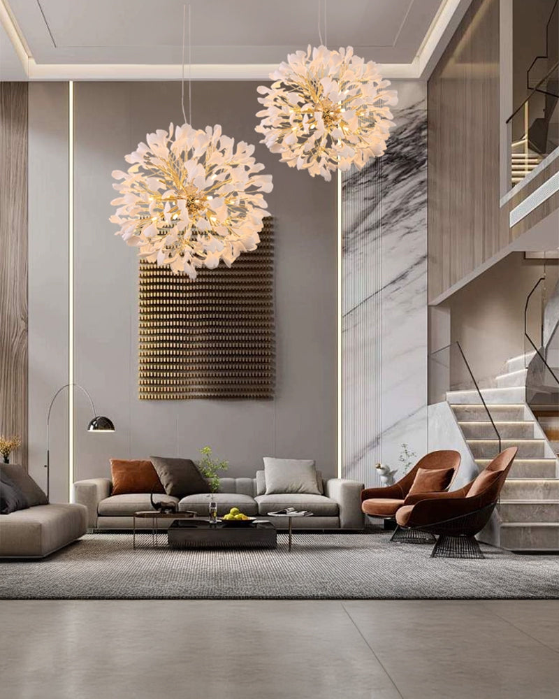 A luxurious living room with neutral decor and a high ceiling, illuminated by two large white and gold ginkgo leaf chandeliers hanging over a gray sectional sofa.