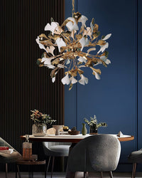 Close-up of a modern dining area with a wooden table and gray chairs; a detailed view of a white and gold ginkgo leaf chandelier suspended above the table is highlighted.