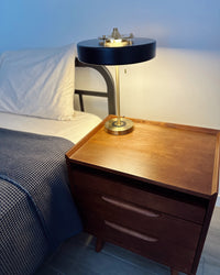 Black bedside lamp glowing on a wooden nightstand beside white bedding and a dark textured blanket.