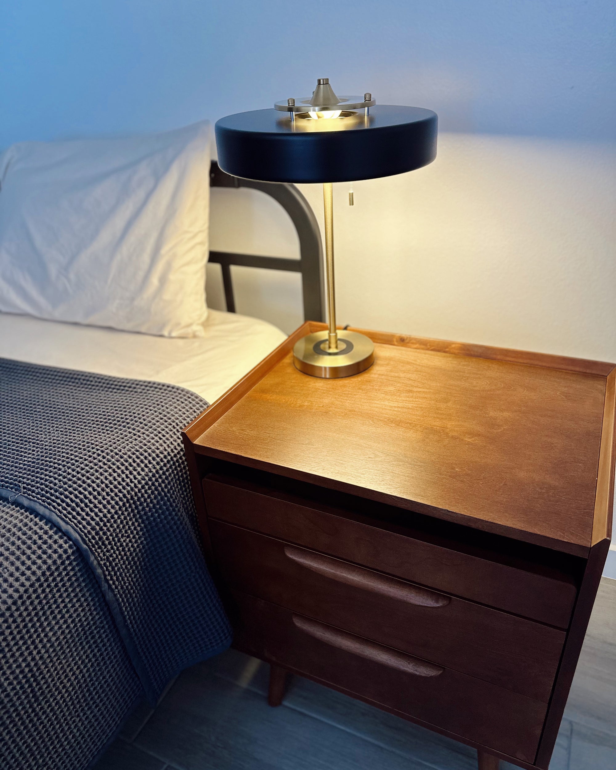 Black bedside lamp glowing on a wooden nightstand beside white bedding and a dark textured blanket.