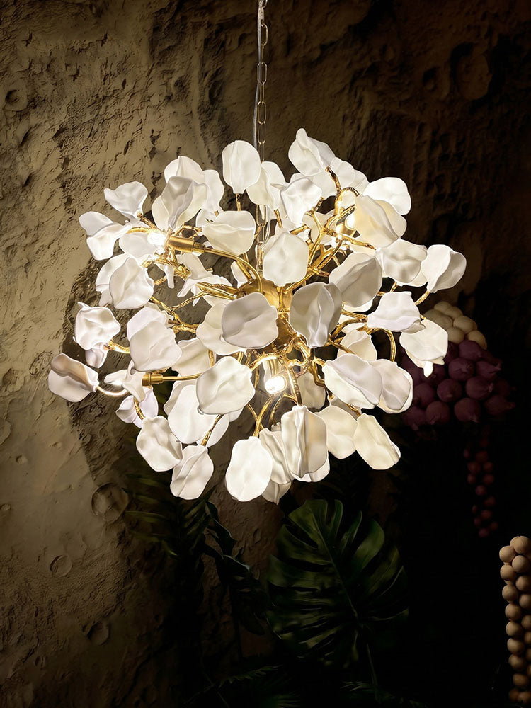Illuminated Art Deco chandelier with layered white ceramic leaf-and-petal forms on golden brass arms against a textured wall and dark greenery.