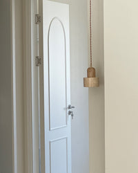 A natural pendant light with a smooth travertine cylinder and light wood top hanging beside a white doorway, shown unlit.
