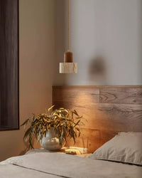 A minimalist pendant light with a smooth travertine shade and dark walnut top, casting a warm pool of light beside the bed.
