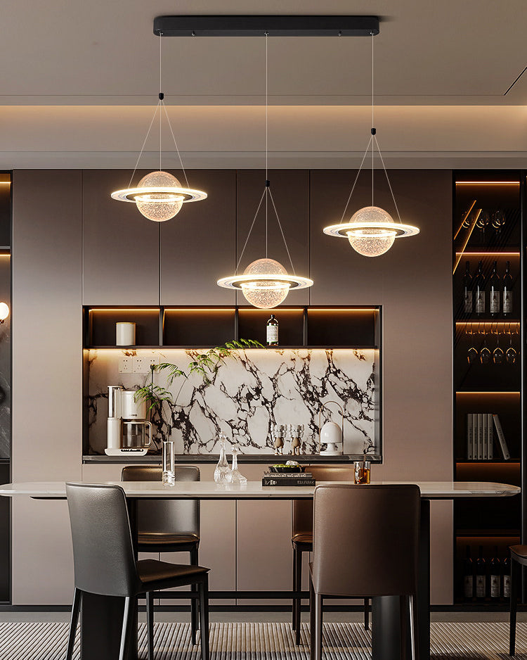 Illuminated modern linear chandelier with three textured planet globes and LED orbit rings above a dining counter with a marble backsplash.