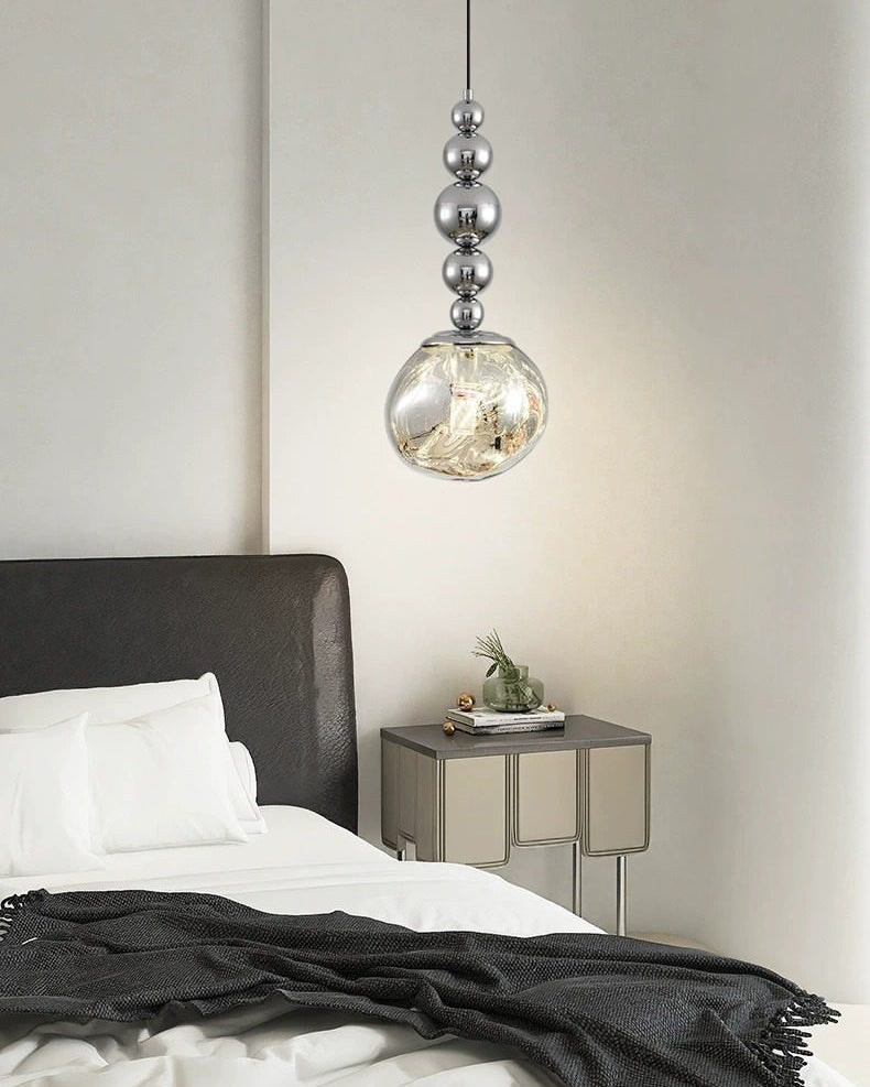 Silver pendant light with five mirrored spheres and an irregular acrylic globe above a metal-trimmed nightstand beside a black-and-white bed.