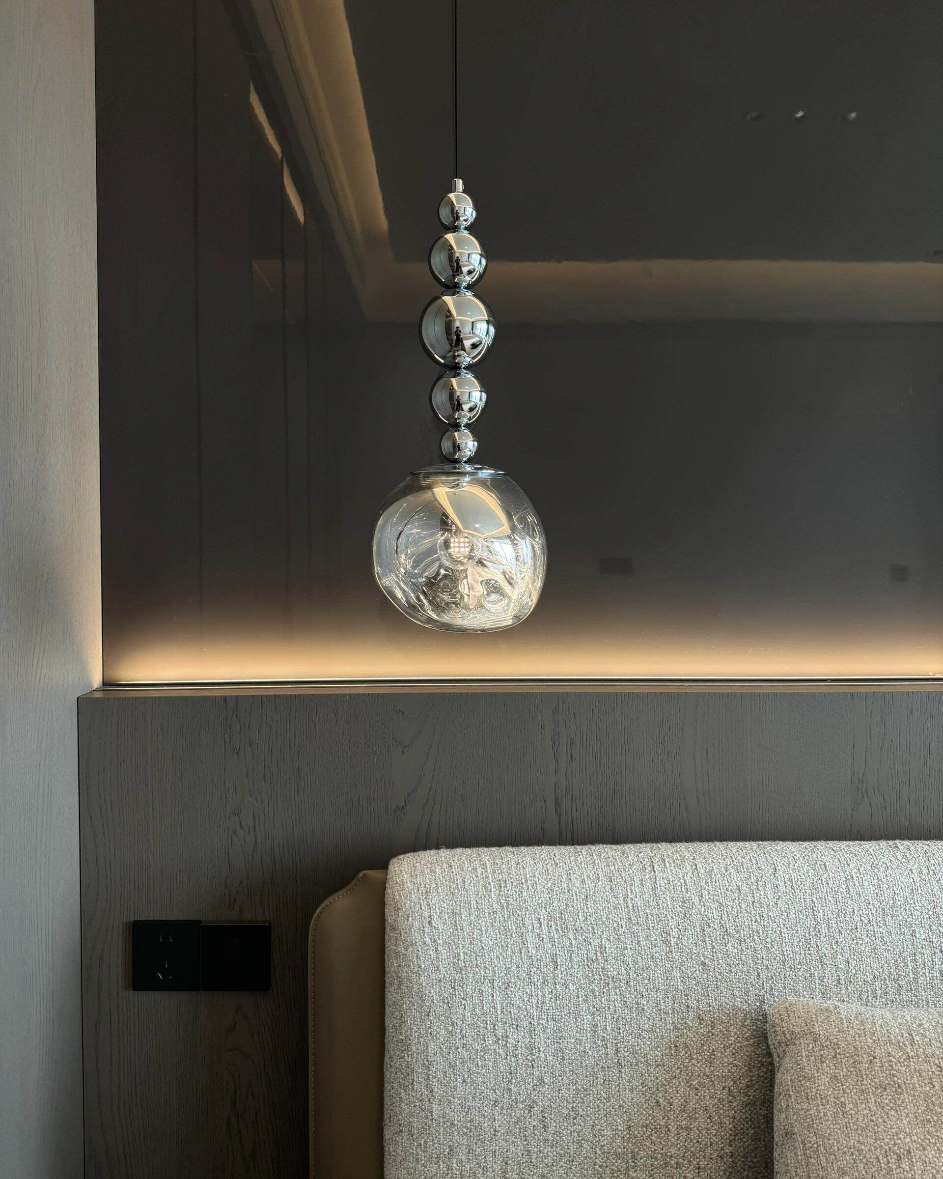 Modern globe pendant light with five mirrored metal spheres and a rippled acrylic globe above a light upholstered headboard against dark wall panels.