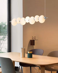 White linear chandelier dining room view with seven glowing glass rings suspended above a pale wood table beside a floor-to-ceiling window.