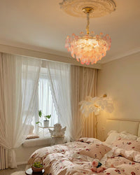 Pink glass flower pendant light with layered ribbed petals above floral bedding and softly draped white curtains.