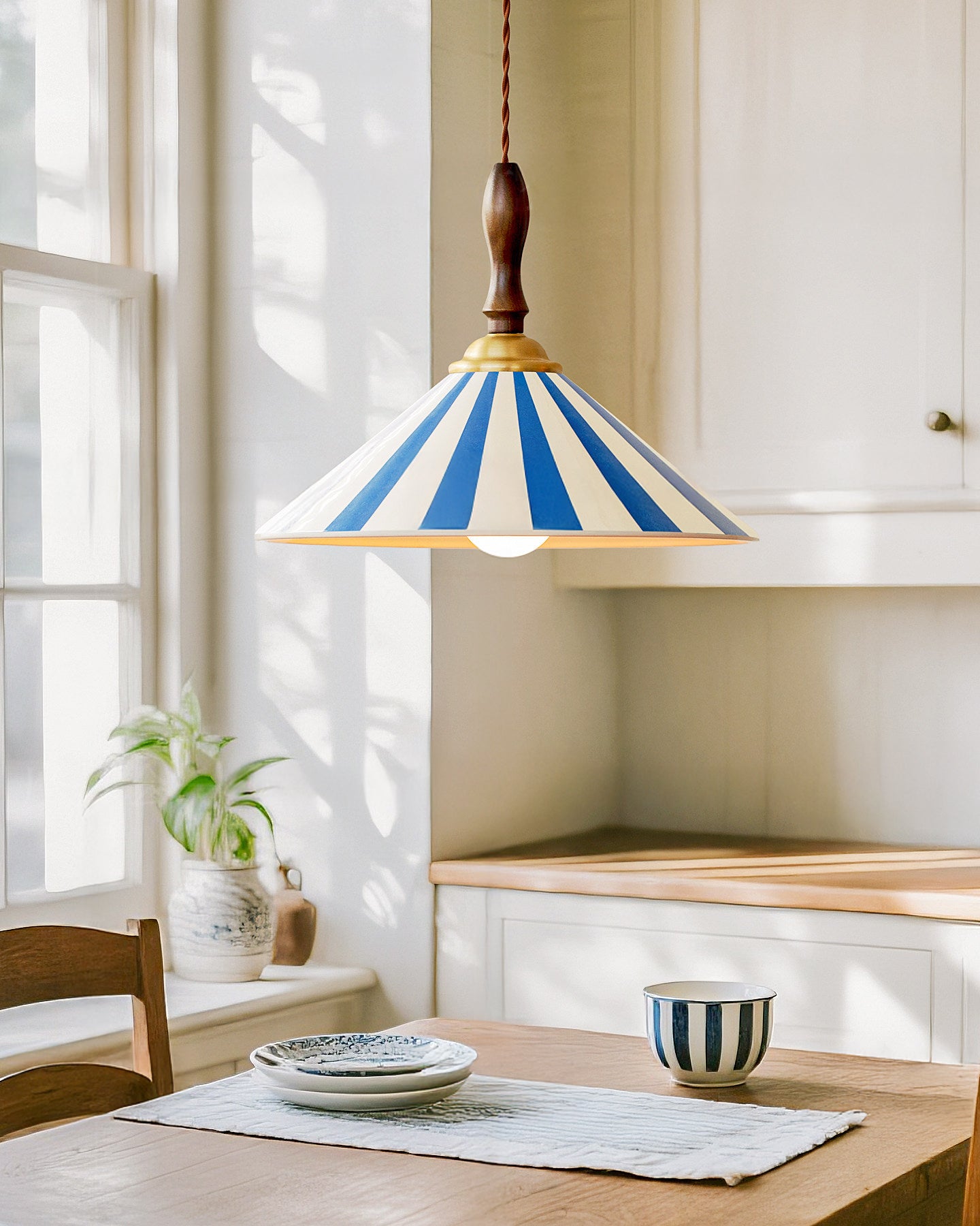 Blue-striped cone pendant light hanging above a wooden kitchen table beside a sunlit window, white cabinetry and striped tableware.