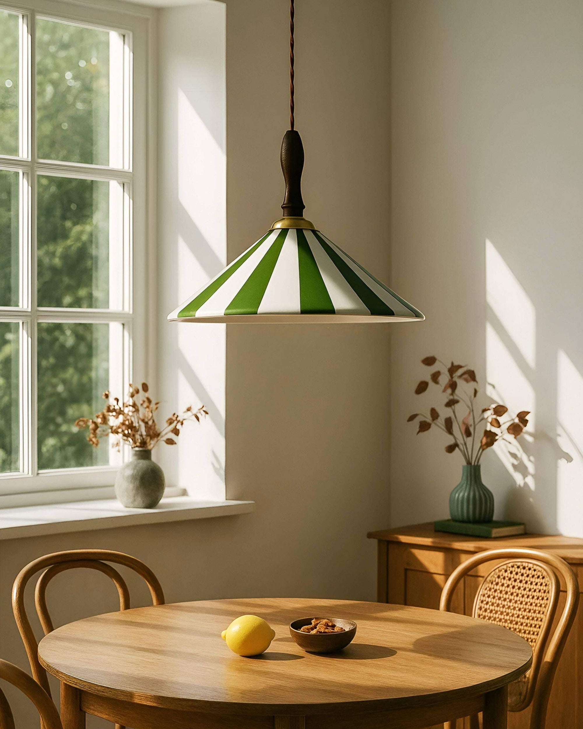 Green-and-white suspended ceiling lamp above a round wood dining table in a sunlit room with cane chairs, framed art and potted plants.
