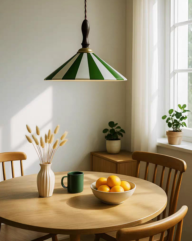 Green-striped cone pendant light fixture above a round breakfast table with oranges, a green mug and natural wood furniture beside a tall window.