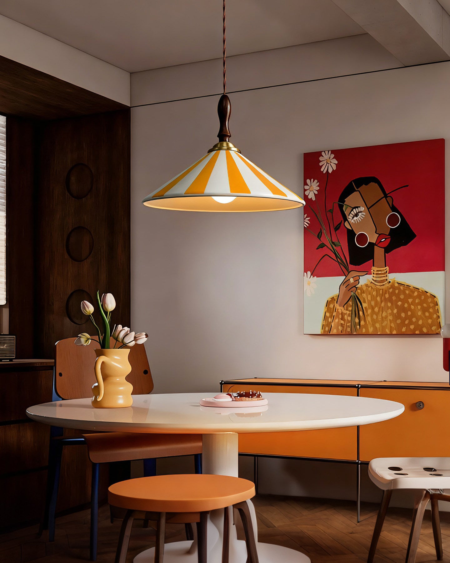 Yellow-striped ceramic fixture showing retro pendant lighting above a white round table in a colorful dining room with orange seating and bold portrait art.
