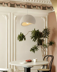 Cream eggshell dining light fixture above a round table, framed by decorative wall molding, indoor greenery, and a soft warm glow.