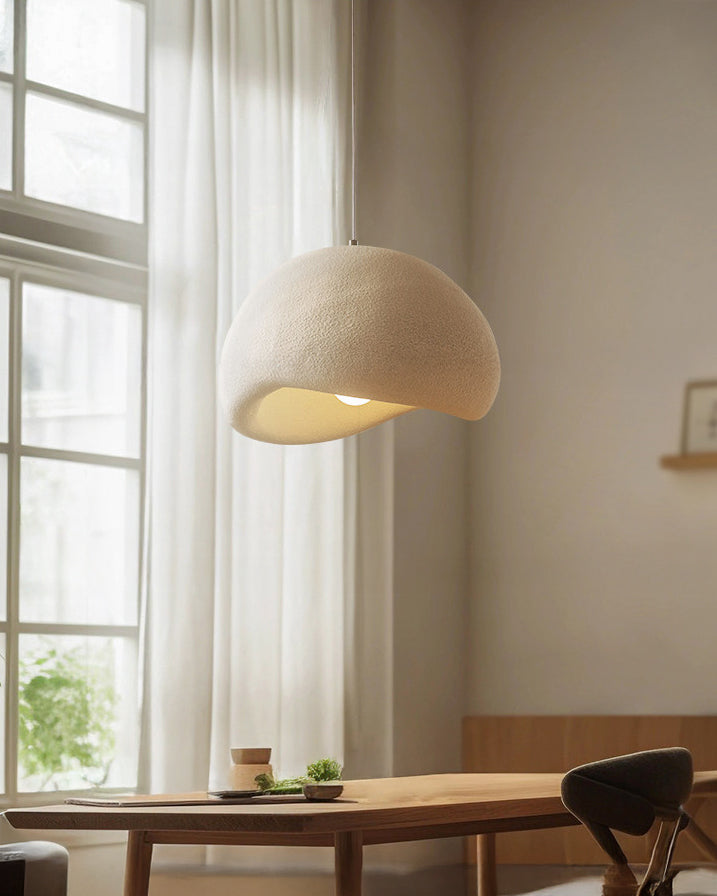 Close view of an organic modern pendant light with a cream textured eggshell shade glowing above a wooden table beside tall windows.