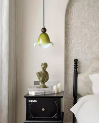 Olive glass hanging lamp glowing softly above a black bedside cabinet in a quiet bedroom with pale botanical wallpaper.