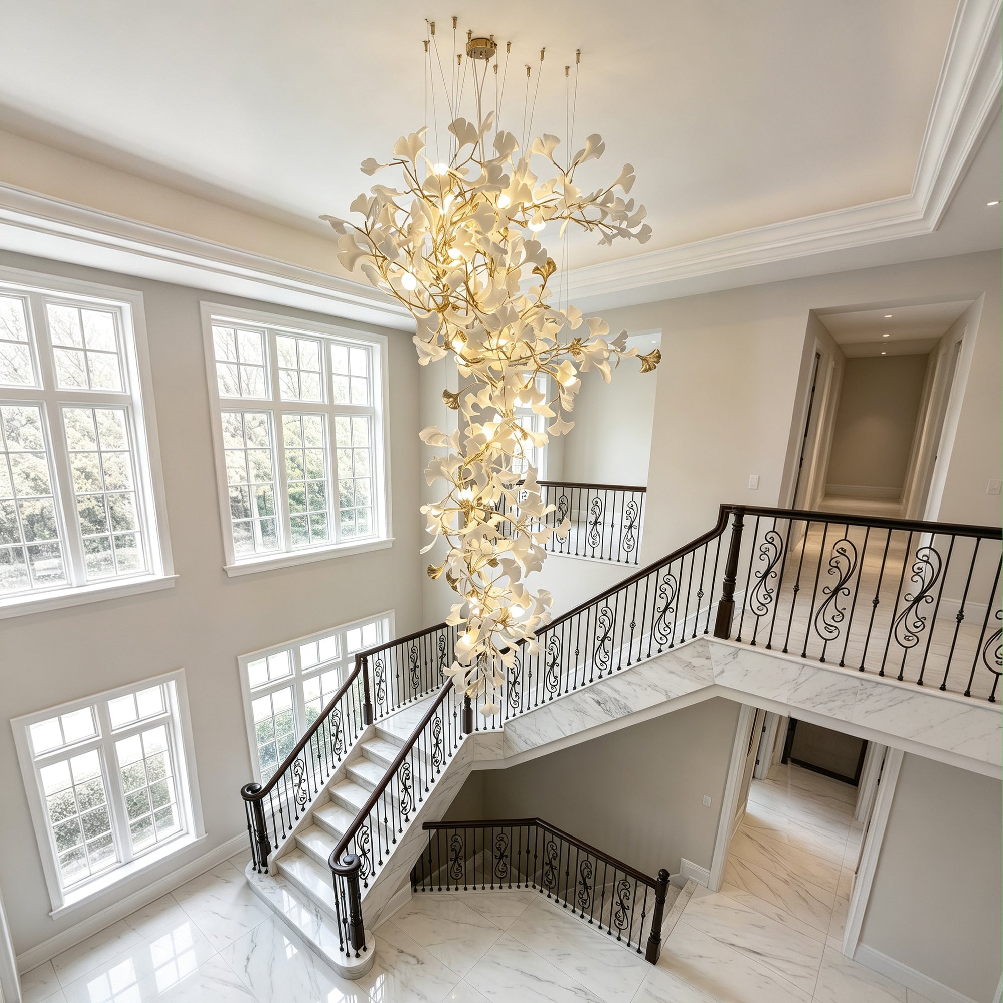 Staircase chandelier with white ceramic ginkgo leaves and gold-toned branches viewed from an upper landing in a bright curved stairwell.