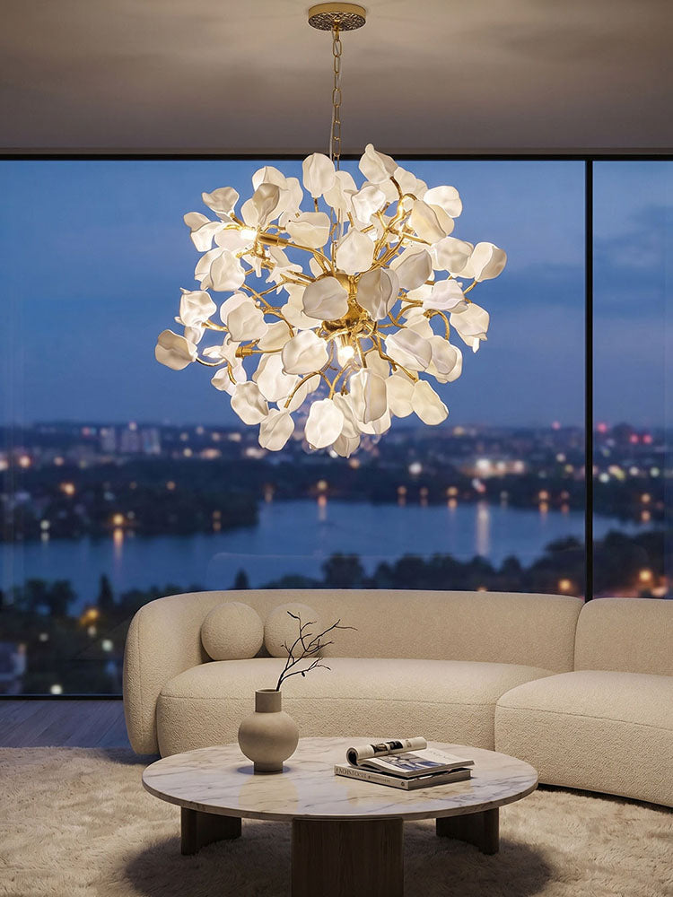 Illuminated white chandelier with ceramic ginkgo-like leaves and golden branches above a cream sofa, framed by windows and an evening city view.