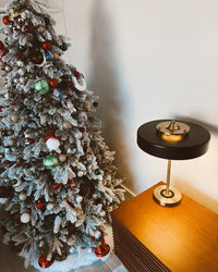 Black mid century table lamp glowing on a wooden cabinet beside a snow-dusted decorated Christmas tree.
