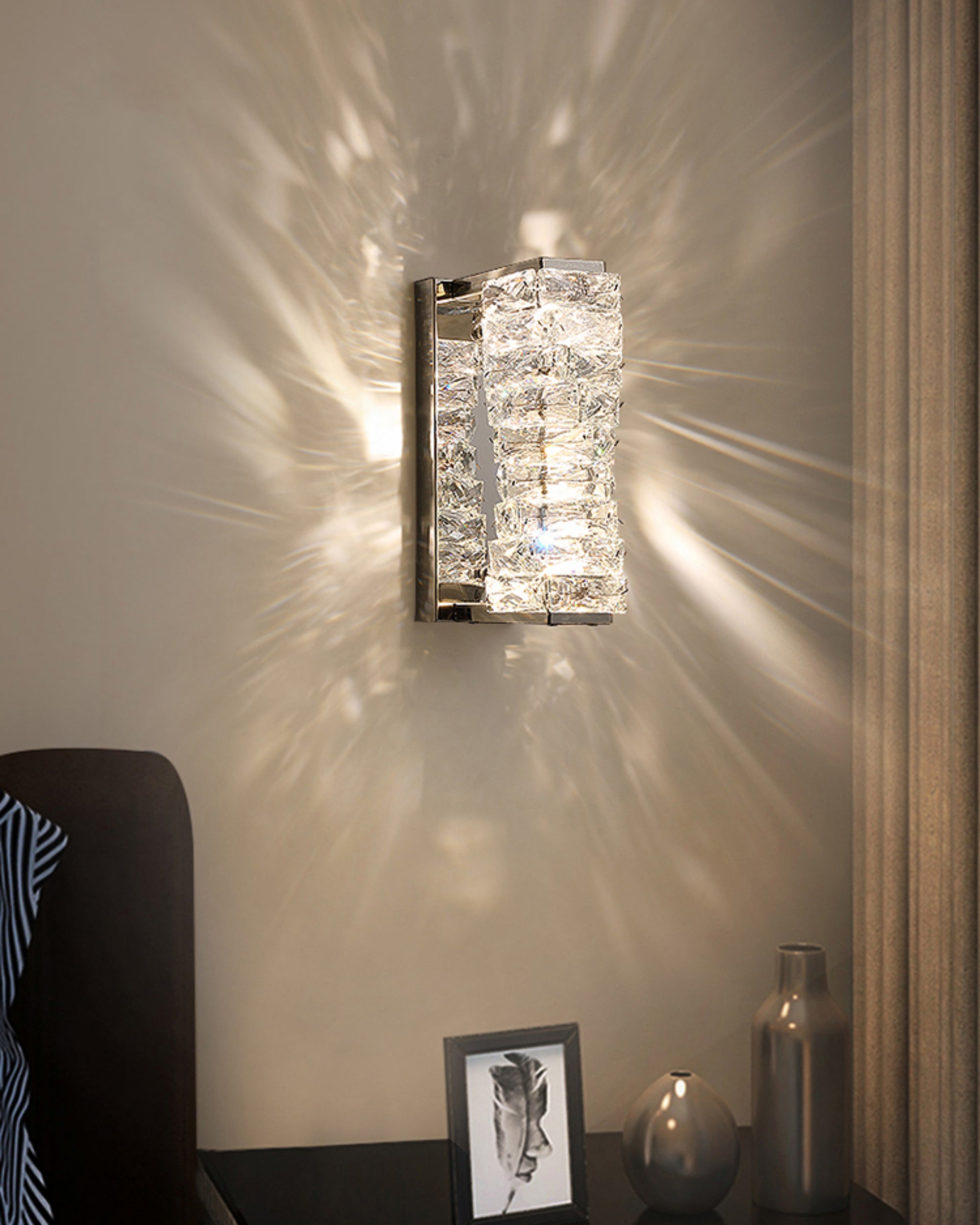 Vertical crystal wall sconce in chrome finish lit on a neutral wall, showing a prismatic light pattern