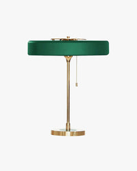 Green mid century table lamp with a wide round metal disc shade, slim gold-tone frame, circular base and hanging pull chain on white.