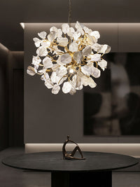 Illuminated gold chandelier with white ceramic leaf forms and branching brass arms above a round black table in a dark modern interior.