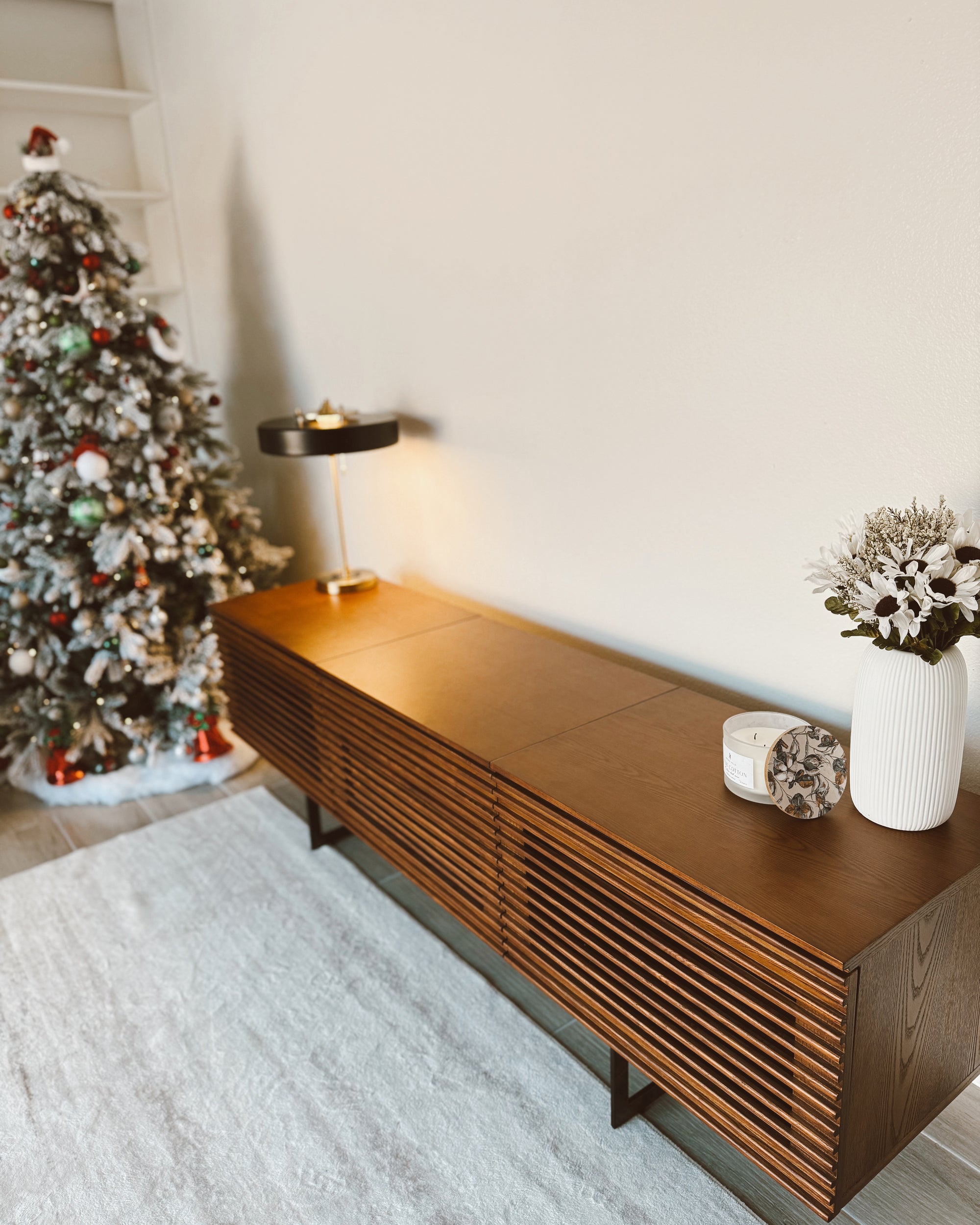 Black vintage table lamp casting warm light from a long wooden console beside a decorated Christmas tree and white vase.