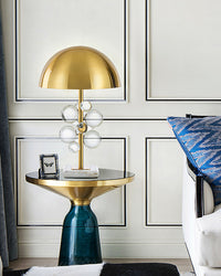 Clear brass lamp with acrylic spheres and a marble base on a blue glass side table beside a white chair.