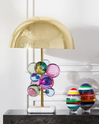 Colorful sculptural table lamp displayed on a black console beside two striped glass ornaments in a bright white interior.