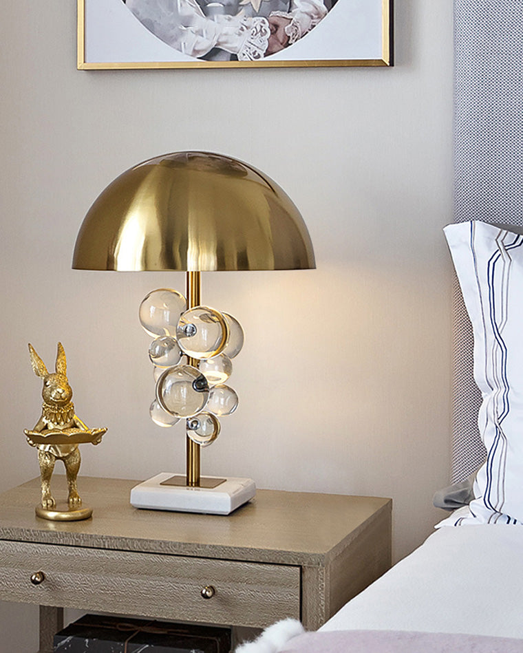 Clear marble table lamp glowing on a neutral bedroom nightstand beside a gold rabbit figure and white bed.