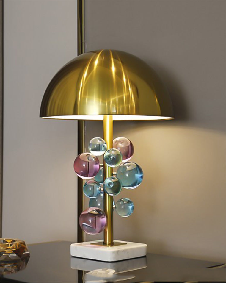 Illuminated gold dome table lamp with jewel-toned acrylic orbs and a white marble base on a reflective black console.