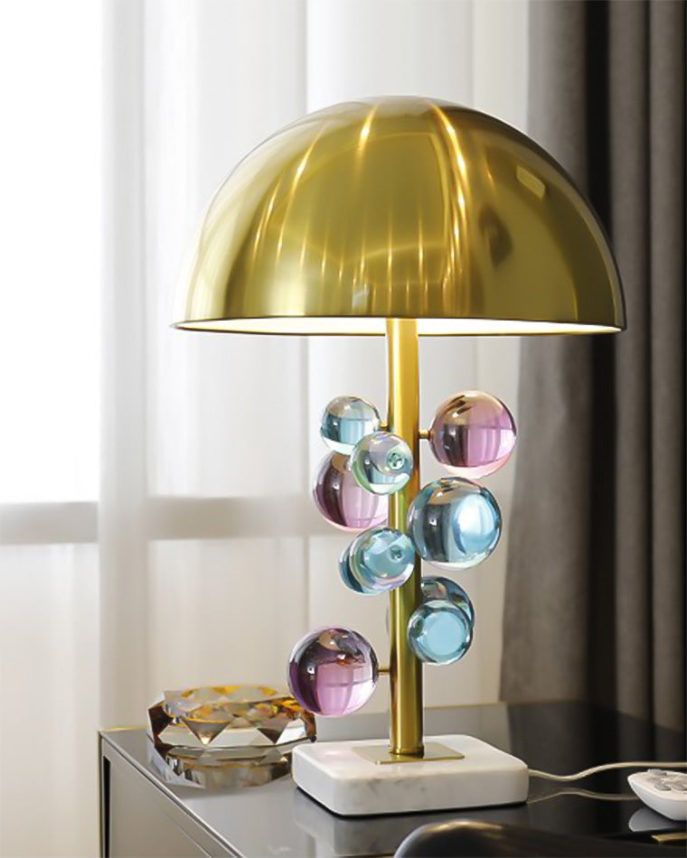 Colorful sculptural table lamp on a black desk by a window, showing the brass dome and layered acrylic spheres.